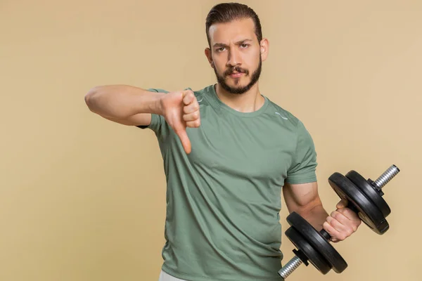 Upset tired athlete sportive man showing thumbs down sign gesture, expressing discontent, disapproval, dissatisfied, dislike. Middle eastern sportsman guy weightlifting dumbbell on beige background
