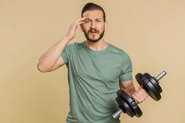 Athlete sportive man showing indignant expression, asking why, what reason of failure, demonstrating disbelief irritation by troubles. Middle eastern sportsman guy with dumbbell on beige background
