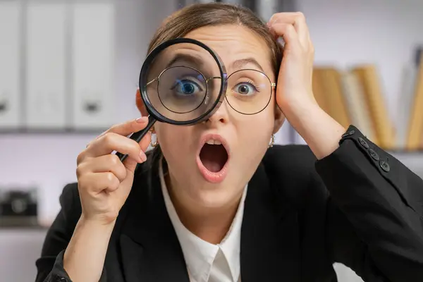 stock image Investigator researcher scientist business woman working at office holding magnifying glass near face looking into camera with big zoomed funny eye, searching, analysing. Professional freelancer girl