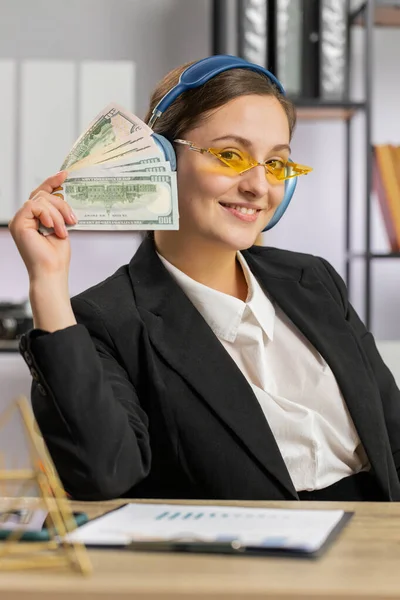 Business woman holding fan of cash money dollar banknotes celebrate, dance, success career, lottery jackpot game winner, big income, wealth at office workplace desk. Remote distant working. Vertical