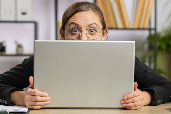 一个自信的女商人躲在笔记本电脑后面 看着摄像机 监视着他的同事们 窥视着 专业的自由职业者 从计算机后面看 眼神狡猾 — 图库照片
