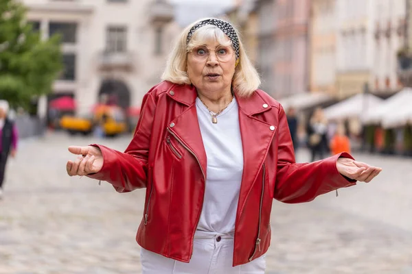What. Why. Surprised frustrated senior woman raising hands asking reason of failure, demonstrating disbelief irritation by troubles outdoors. Confused elderly grandmother walking in urban city street