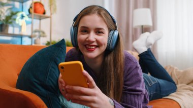 Kablosuz kulaklıklı mutlu genç bir kadın evinde koltukta uzanmış en sevilen enerjik disko dans müziğini dinliyor. İnsanlar hafta sonu boş zaman aktiviteleri