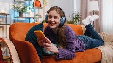 Kablosuz kulaklıklı mutlu genç bir kadın evinde koltukta uzanmış en sevilen enerjik disko dans müziğini dinliyor. İnsanlar hafta sonu boş zaman aktiviteleri