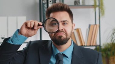 Investigator researcher scientist businessman working at home office holding magnifying glass near face looking into camera with big zoomed funny eye, searching, analysing. Professional freelancer man