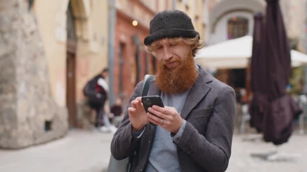 インターネットを閲覧し 仕事を終え カメラを見ているスマートフォンの入力テキストメッセージを使用して若い男を笑顔 赤毛の髭の男が都会の通りの背景を歩いている 町のライフスタイル屋外 — ストック動画