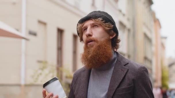 年轻的留着胡子的红头发男子喜欢在户外喝早茶或热茶 休息一下 红头发的家伙走在市中心的街道上 喝咖啡去了 外面的城镇生活方式 — 图库视频影像