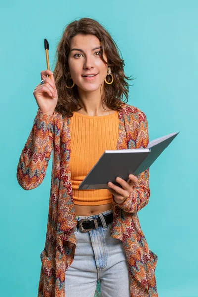 Thoughtful journalist woman making notes, writing down thoughts with pen into notepad notebook diary, to do list, good idea. Pretty brunette student girl isolated on blue studio background. Vertical