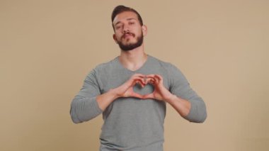 Ben de seni seviyorum. Gülümseyen mutlu Lübnanlı genç adam kalp hareketi yapıyor. Aşk işareti güzel romantik duygular ve sempati sembolü ifade ediyor. Orta Doğulu adam bej renkli stüdyo arka planında izole edilmiş.