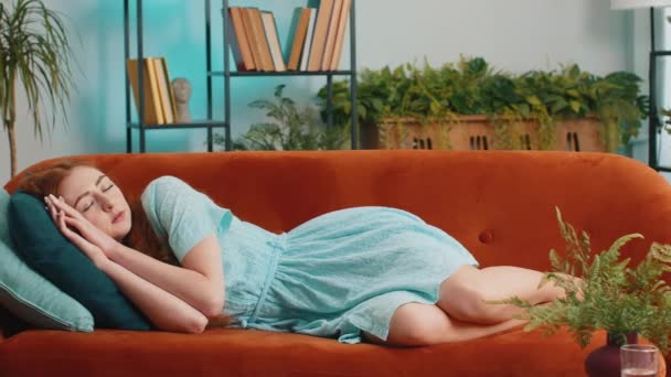 疲れた若い赤毛の女性が家で休むためにベッドに横になっています 屈託のない少女の肖像画 枕と快適なオレンジのソファの上で眠りに落ちる 彼女の目を閉じて一人で昼寝を楽しむ — ストック動画
