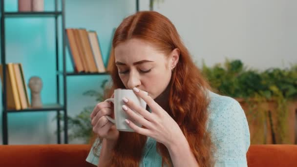 Привлекательная улыбающаяся молодая рыжая женщина пьет чашку теплого кофе или травяной чай, сидя по утрам на диване в гостиной. Любимая девушка, наслаждающаяся комфортным отдыхом, отдыхая одна