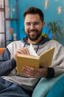 Genç beyaz adam ilginç bir kitabı rahatlatıyor, sayfaları çeviriyor edebiyattan zevk alıyor, rahat bir kanepeye uzanıyor. Huzurlu, neşeli bir adamın portresi. Oturma odasında, kanepede.
