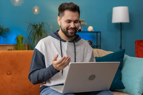 Caucasian man wearing headset, freelance worker, call center or support service operator helpline, having talk with client or colleague communication support waving hello. Guy at home room on sofa