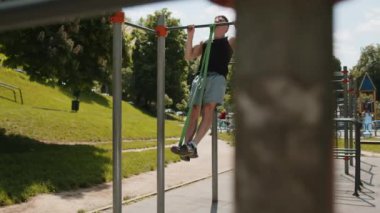 Spor giyimli atletik bir adam elastik genişleticinin yardımıyla barfiks egzersizi yapıyor. Çocuk parkında sırt kaslarını şişiren genç adam. Spor sağlığı rutin egzersizi. Vücut geliştirme