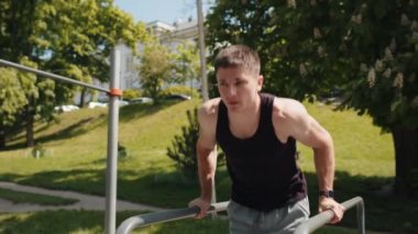 Athletic caucasian man in sportswear practicing dips on parallel bars, training triceps. Young guy in t-shirt on playground. Sports health fitness routine, workout. Strength motivation. Outdoor gym