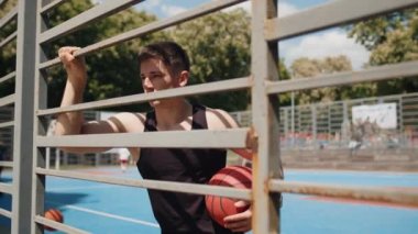 Basketbol oynayan formda beyaz bir adamın portresi. Tek başına poz veren genç, ciddi bir sporcu şehir parkındaki sahada ızgaranın arkasında duruyor. Spor rutini. Motivasyon. Açık havada.