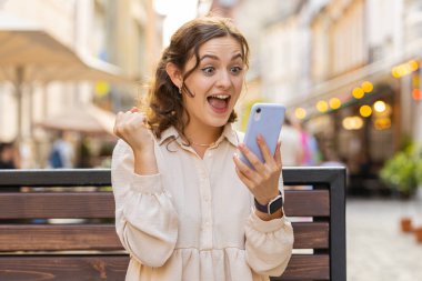 Şaşırmış genç bir kadın cep telefonuyla kutlama yapıyor. İyi haber kazan, piyango kazan, internetten bağış yap. Mutlu güzel kız şehrin güneşli sokaklarında bankta oturuyor. Yaşam Tarzları