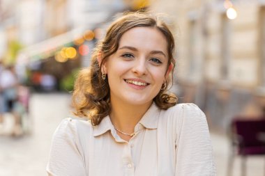 Mutlu güzel bir genç kadının portresi. Dostça gülümseyen, kameraya bakan mutlu yüz ifadesi. Hayal kuran, dinlenen, rahatlayan, dışarıda iyi haberler veren. Şehir Güneşi Sokağı arka planında bir kız