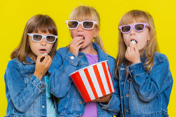 Teenage girls wearing 3D glasses eating popcorn watching interesting tv serial, sport game, film, online social media movie content. Little children sisters. Three siblings kids on yellow background
