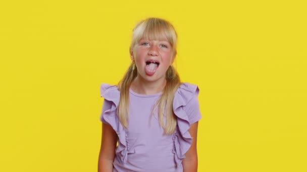 陽気で面白い若いブロンドの女子高生は カメラで顔を作る舌を示し 周りをだます 冗談を言い 愚かな顔をして いじめ 虐待をからかっています 前ティーン女性子供子供上の黄色の背景 — ストック動画