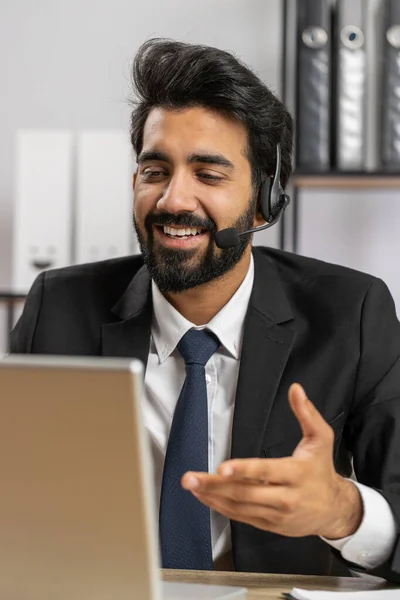 Indian business man working on laptop wearing headset, freelance worker call center or support service operator helpline, having pleasant talk with client or colleague communication support at office