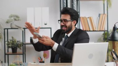 Neşeli genç Hintli iş adamı dizüstü bilgisayarla çalışıyor ve kameraya para harcıyor. Daha fazla bahşiş, daha fazla kar getirir. Piyango alışverişi kazanır. Ev ofis masasındaki Bağımsız Arap Adam 'ın kutlu olsun.