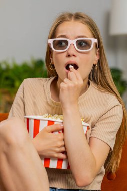 Kanepede oturup patlamış mısır yiyen ve ilginç TV dizileri, spor oyunları, filmler, evdeki sosyal medya içerikleri izleyen heyecanlı genç bir kadın. Üç boyutlu gözlüklü kız ev eğlencesi yapıyor.