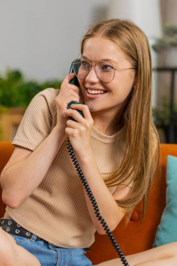 Genç sarışın bir kadın arkadaşlarıyla telefon görüşmesi yapıyor. Mutlu, heyecanlı yetişkin bir kız, 80 'lerden kalma eski moda telefonlardan keyif alıyor. Oturma odasında konuşuyor. Turuncu koltukta oturuyor.