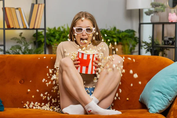 Excited young woman sitting on sofa eating popcorn watching interesting horror TV film movie online social media content serial, sport game at home. Scared girl in 3D glasses spilling popcorn on couch