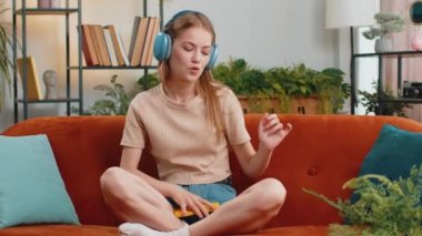 Portrait of happy overjoyed woman in wireless headphones relaxing sits on couch at home apartment choosing listening favorite energetic disco dancing music. People weekend daytime leisure activities