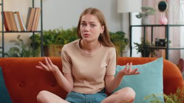 I dont know what to say. Confused pretty woman feeling embarrassed about ambiguous question, having doubts, no idea, being clueless and uncertain at home. Portrait of girl sits on sofa in living room