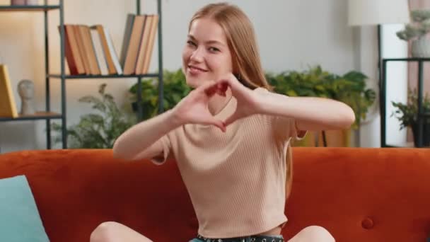愛してるわ 家庭のリビングルームのソファで幸せな若い女性の愛の象徴になり カメラに心臓サインを示し ロマンチックな感情を表現誠実な肯定的な感情を表現します — ストック動画