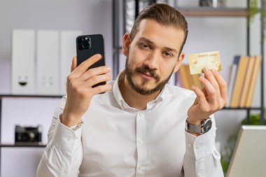 Genç bir ortadoğulu işadamı kredi kartı ve akıllı telefon kullanıyor. İnternetten alışveriş siparişleri alıyor. İş yerindeki serbest çalışan adam kameraya bakıyor.
