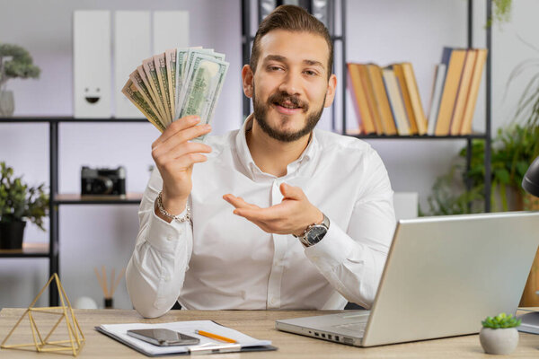 Young middle eastern boss business man showing money dollar cash gives to you wages salary, satisfied with good work. Success, wealth, investment. Freelancer guy at office workplace looking at camera