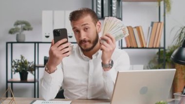 Akıllı telefon gösteren iş adamı içten bir sevinçle kazanıyor, nakit para alıyor, piyango başarısı, telefonla kazanmayı öneriyor. İş yerindeki serbest çalışan adam kameraya bakıyor.