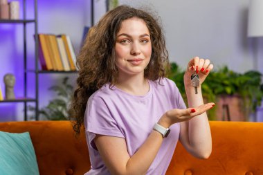 Esmer kadın emlakçı el kaldırıyor. Yeni ev dairesinin anahtarlarını gösteriyor. Ev satın alıyor ya da kiralıyor. Odadaki kız turuncu koltukta oturuyor.