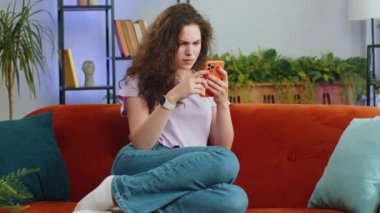 Hüzünlü kadın akıllı telefon daktilo kullanıyor, beklenmedik piyango sonuçlarını kaybediyor, kötü haber, servet kaybı, başarısız teslim tarihi, virüs. Oturma odasında kanepede oturan genç kızın portresi.