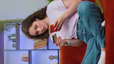 Kredi kartı ve akıllı telefon kullanan genç bir kadın çevrimiçi alışveriş nakitlerini naklederken ev içinde yemek siparişi veriyor. Mutlu kız kanepede oturuyor. Dikey