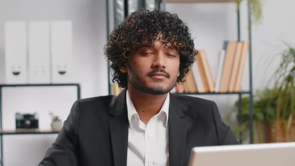 現代のオフィスの職場の机で幸せな笑顔ひげを生やしたインドのビジネスマンの肖像画離れてカメラで頭を向ける見てください 正式なスーツを着た男の後ろに ハンサムな男 遠隔地のオンライン作業ノートパソコン上で — ストック動画
