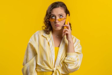 Gözlüklü, uykulu genç bir kadın arkadaşıyla telefonda konuşuyor. İnternette sohbet ediyor. Sarı arka planda izole edilmiş akıllı telefondan sıkıcı konuşmalar yapan ilgisiz çekici bir kız.