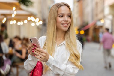 Gezgin, sarışın, akıllı telefon kullanarak açık hava mobil navigatör uygulamasında haritada arama yapmak için kısa mesajlar yazan genç bir kadın. Kentin arka sokaklarından geçen genç bir kız.