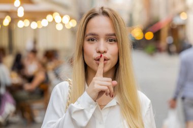 Sessiz olun lütfen. Sarışın genç bir kadın işaret parmağını dudaklarına bastırıyor. Sessiz ol işareti yapıyor. Dışarıdaki dedikoduları kimseye söyleme. Şehir caddesinde yürüyen genç bir kız. Kasaba yaşam tarzları