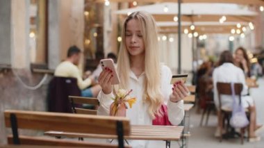 Para transfer ederken kredi kartı akıllı telefonu kullanan genç bir kadın, online alışveriş, yemek siparişi, açık hava otel odası rezervasyonu. Genç kız turist kafeteryada masada oturuyor.