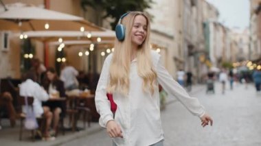 Kablosuz kulaklıklı mutlu sarışın kadın favori enerjik müziği dinliyor, zaferi kutluyor. Genç kız turist şehir caddesinde yürüyor. Şehir yaşam tarzları açık havada