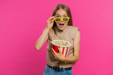Üç boyutlu gözlüklü, patlamış mısır yiyen ve ilginç TV dizileri, spor oyunları, filmler, çevrimiçi sosyal medya filmi içeriği izleyen heyecanlı genç bir kadın. Sarışın, sevimli kız pembe stüdyo arka planında izole edilmiş.