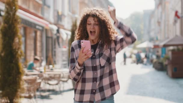 かなり若い女性は 良いメッセージニュース 宝くじのジャックポットの勝利 プレゼントをオンラインで屋外で受賞を祝う携帯電話のスマートフォンを使用しています 都会の日差しの街を歩く幸せな女の子の観光客 町の暮らし — ストック動画