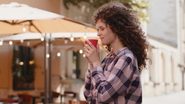 朝のコーヒーのホットドリンクと笑顔を屋外で楽しむ幸せなかなり若い女性 リラックスして休憩 女の子都市部の日差しの通りを歩くと コーヒーを飲みに行く 外の町の生活 — ストック動画
