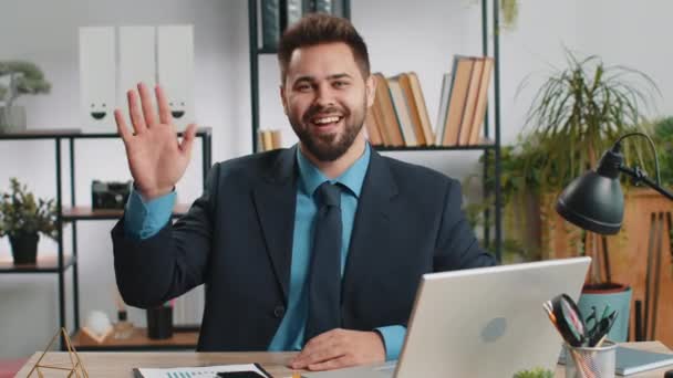 こんにちは カメラで笑顔のラップトップコンピュータで働く白人ビジネスマン 手を振るジェスチャーハイ 挨拶や別れ ホームオフィスの職場デスクでホスピタリティの表現を歓迎 — ストック動画