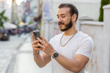 Yakın plan gülümseyen sakallı genç adam akıllı telefon kullanıyor internet sosyal medya uygulamalarını tarıyor, çalışıyor, dışarıda çalışıyor. Şehir Güneşi Şehri caddesinde yürüyen bir turist. Kasaba yaşam tarzları
