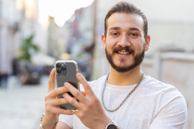 Gülümseyen sakallı genç adam akıllı telefon kullanıyor mesajlar yazıyor internette geziniyor, işi bitiriyor, dışarıdaki kameralara bakıyor. Şehir Güneşi Şehri caddesinde yürüyen bir turist. Kasaba yaşam tarzları